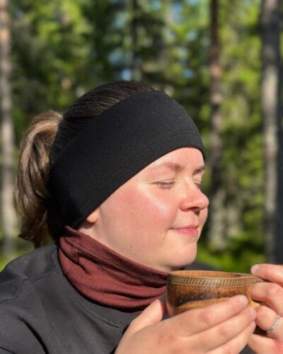 Naisten musta merinovillapanta ja tummanpunainen (rubiininpunainen) tuubihuivi naisella, joka nauttii kahvia ulkona kuksa kädessään.