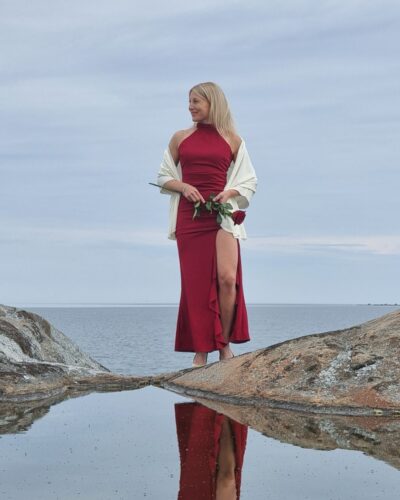Iso luonnonvalkoinen merinovilla kaulahuivi (hartiahuivi). Punaiseen juhlamekkoon pukeutuneen naisen harteilla, rantakalliolla.