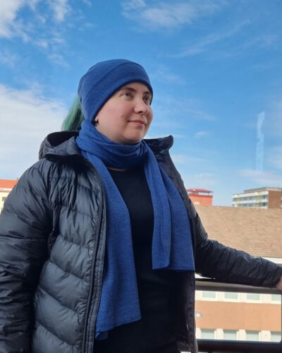 Naisten sininen merinovilla kaulahuivi ja ponnaripipo vihreähiuksisella naisella. Ios kaulahuivi on kiedottu kaulaan ja hiukset on vedetty ponnaripipon aukosta ulos.