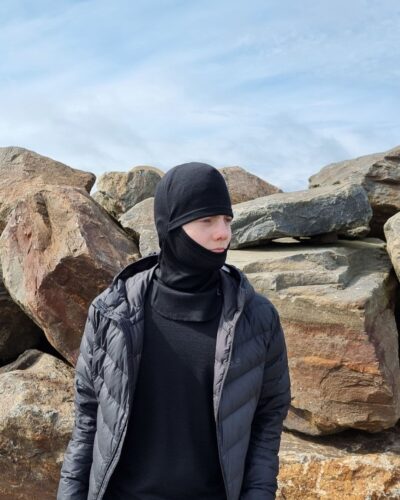 Lasten musta merinovilla kypärämyssy (balaclava). Lasten musta merinovilla kypärämyssy (balaclava). Nuoren miehen päässä etuviistosta kuvattuna.