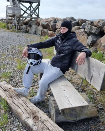 Lasten musta merinovilla kypärämyssy (balaclava). Nuoren miehen päässä joka istuu penkillä mopokypärä sylissään.