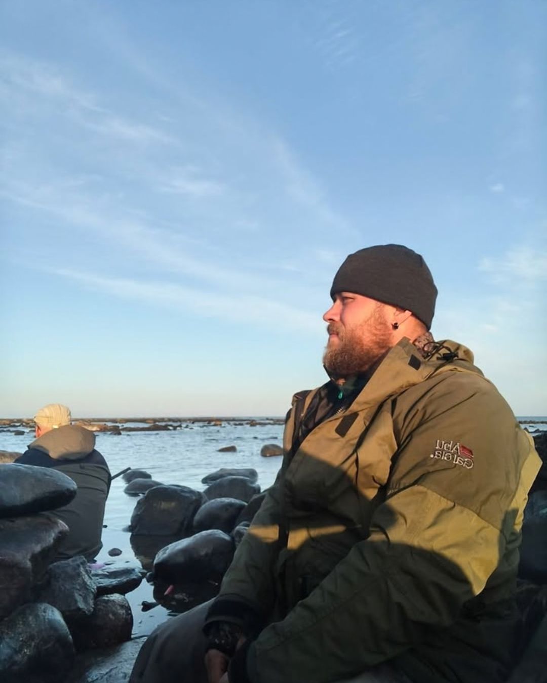 Miesten tummanharmaa merinovillapipo rannalla istuvan metsästäjän päässä. Kuvattu otettu sivusta.