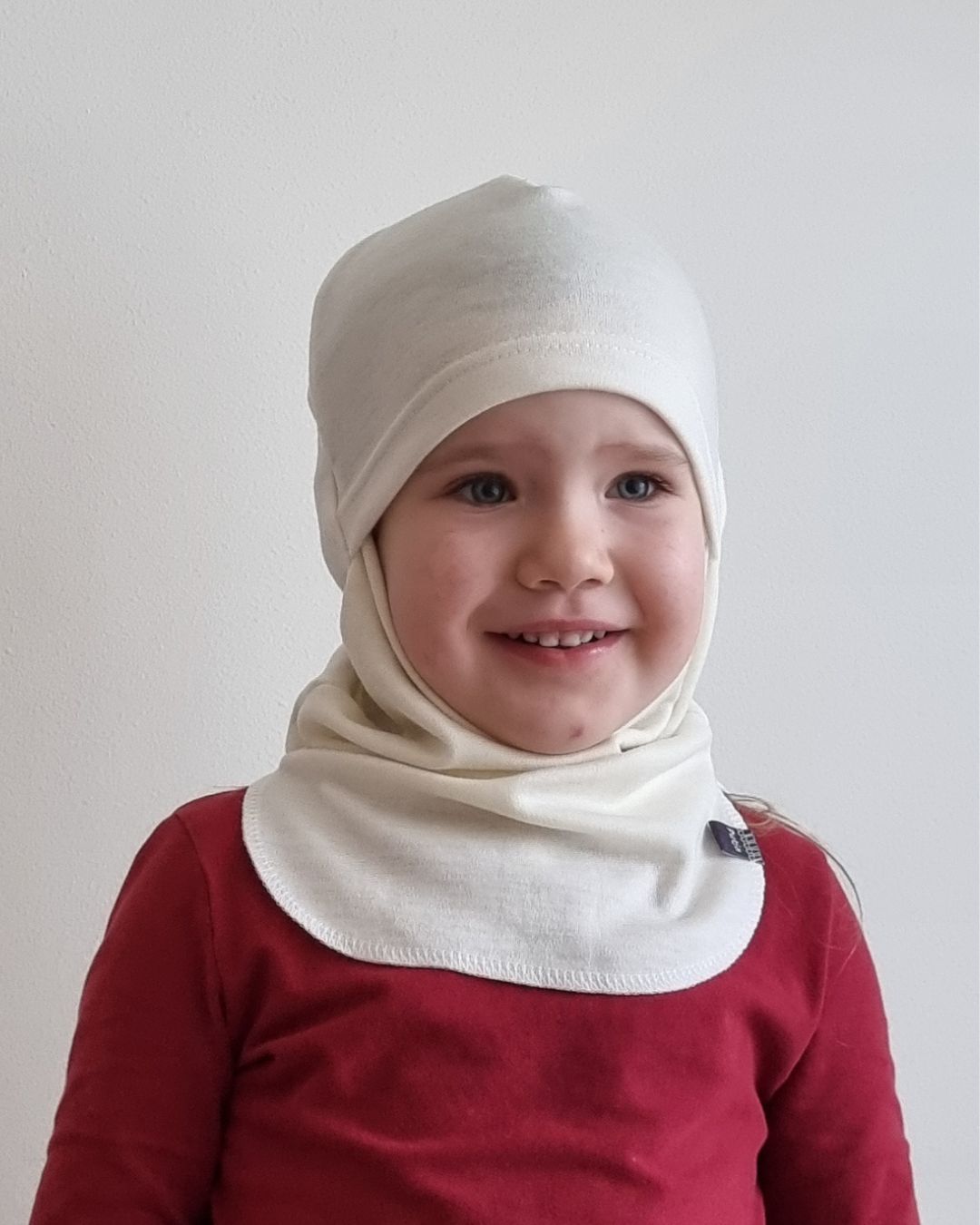 Lasten luonnonvalkoinen merinokypärämyssy kasvosuojalla (kommandopipo) pienen tytön päässä. Lähikuva edestä.