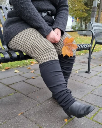 Pitkät mustat merinovilla säärystimet penkillä istuvan naisen jalassa, saappaiden päällä nilkkoihin rypytettyinä.