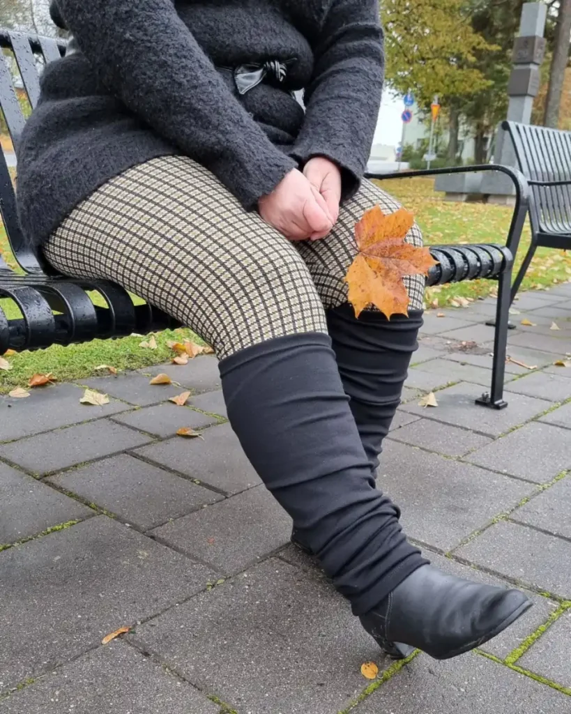 Pitkät mustat merinovilla säärystimet penkillä istuvan naisen jalassa, saappaiden päällä nilkkoihin rypytettyinä.