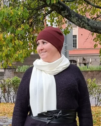 Naisten rubiininpunainen merinovillapipo ja luonnonvalkoinen kaulahuivi naisen päällä syksyisessä kaupunkimaisemassa.