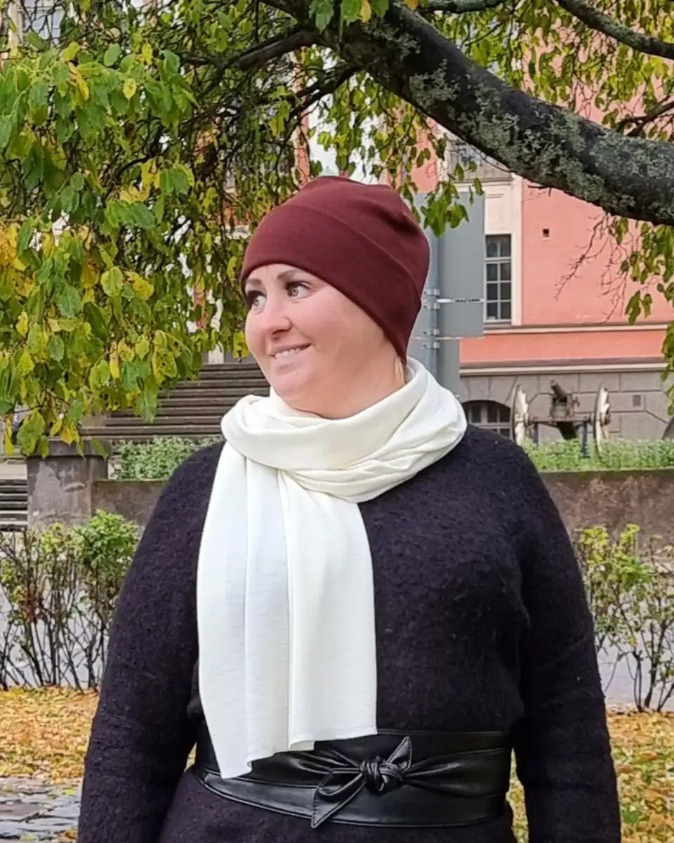 Naisten rubiininpunainen merinovillapipo ja luonnonvalkoinen kaulahuivi naisen päällä syksyisessä kaupunkimaisemassa.